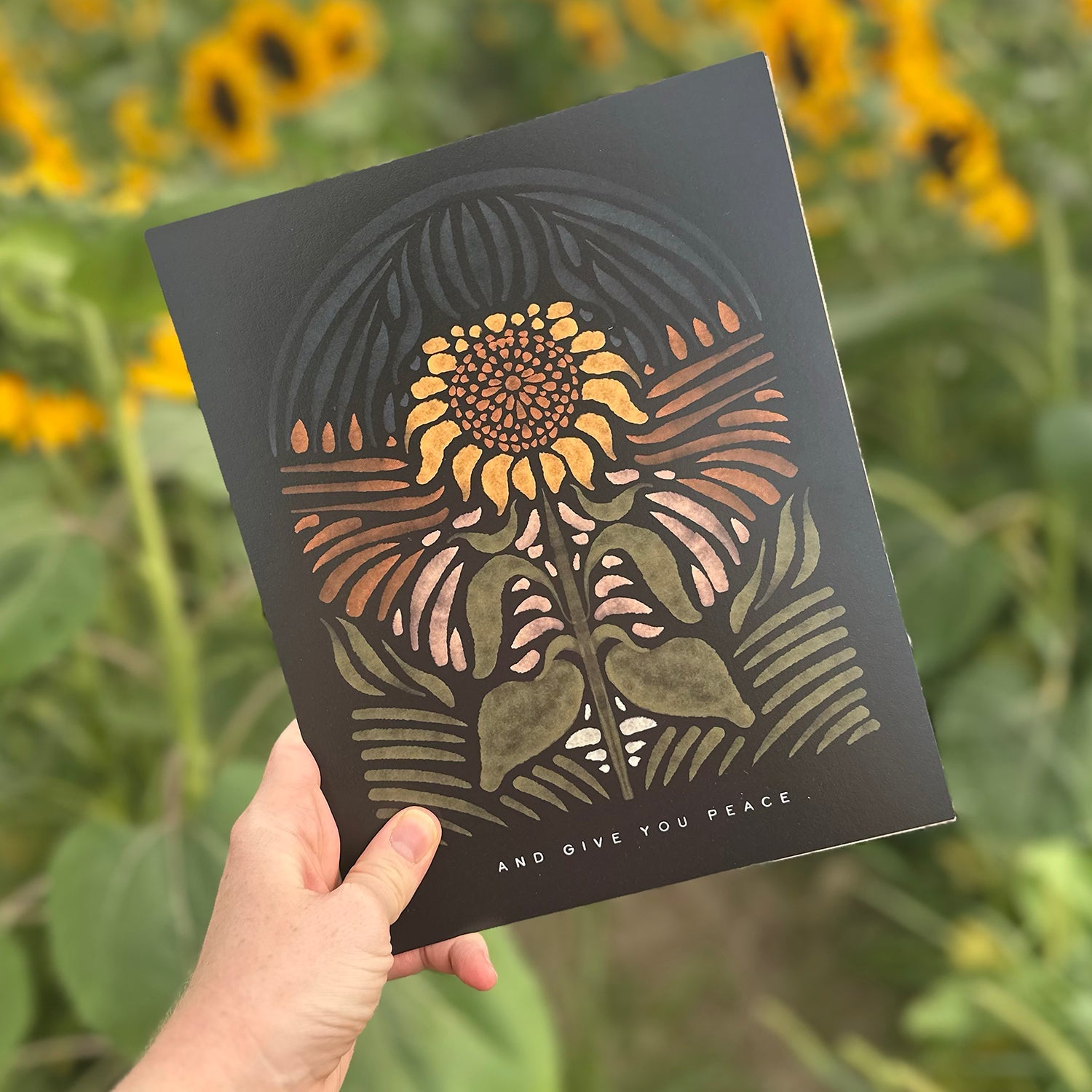 Hand holding art print of a sunflower with it's head up at night with the text "and give you peace"