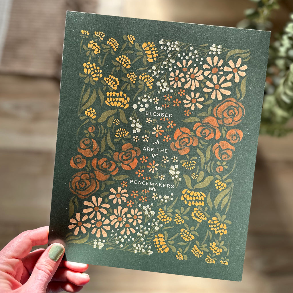 Hand holding art print of floral illustration that surrounds the text "blessed are the peacemakers"