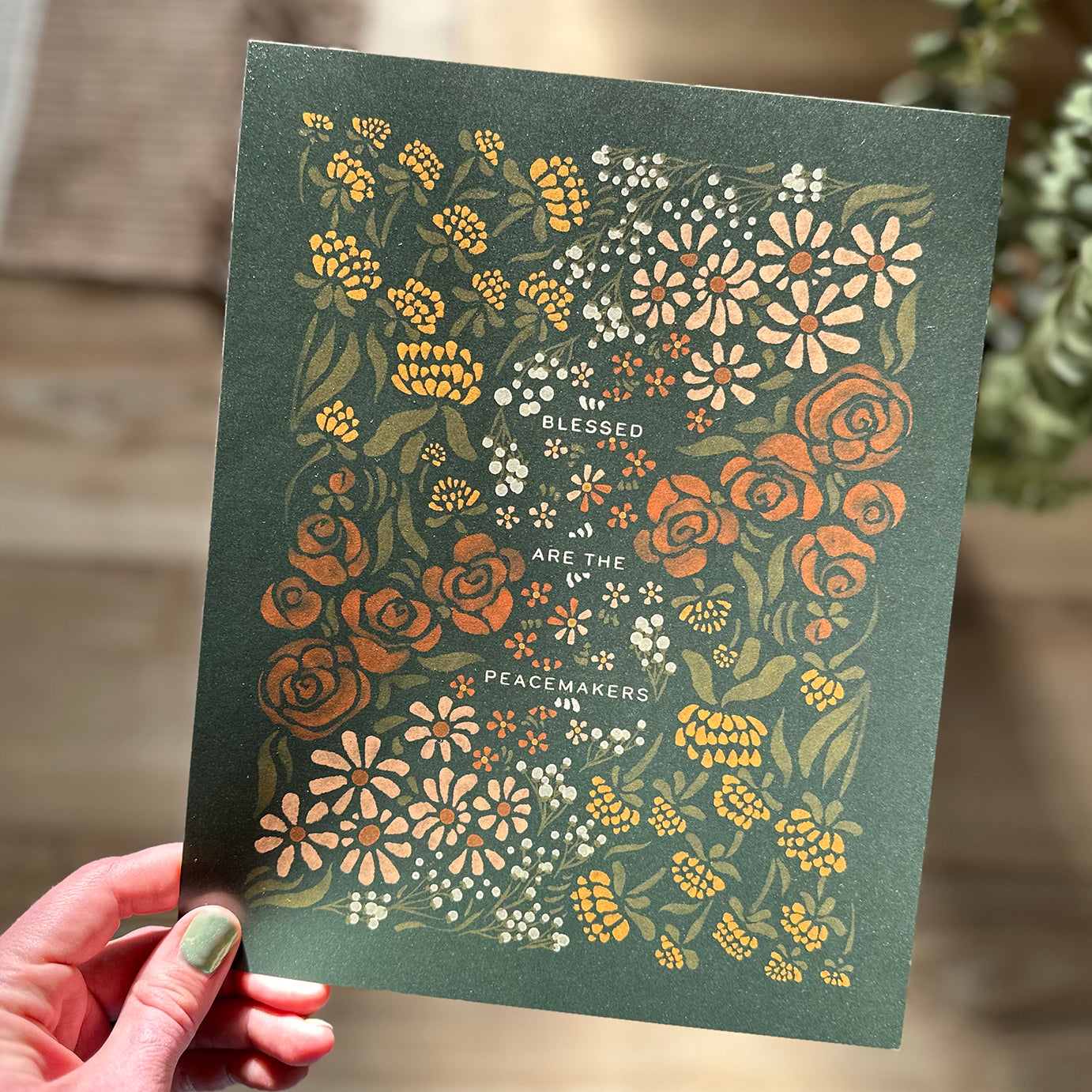 Hand holding art print of floral illustration that surrounds the text "blessed are the peacemakers"