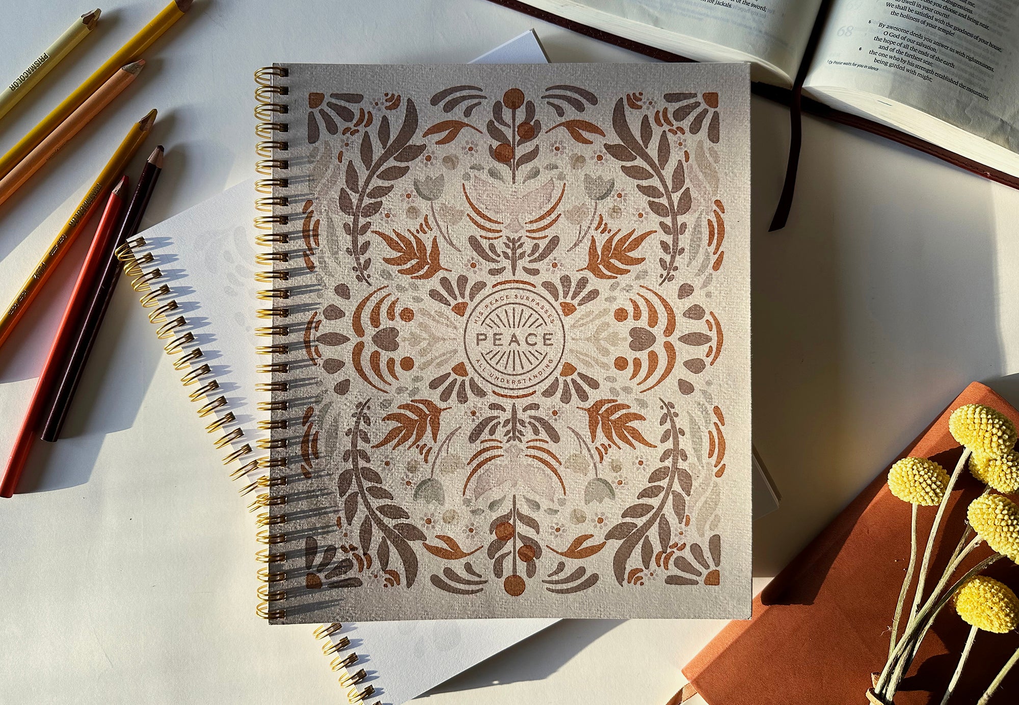 Notebook with decorative cover featuring the word 'Peace', surrounded by pencils and a book on a light surface.