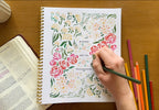 Person coloring a floral design in a spiral-bound notebook with colored pencils on a wooden table.
