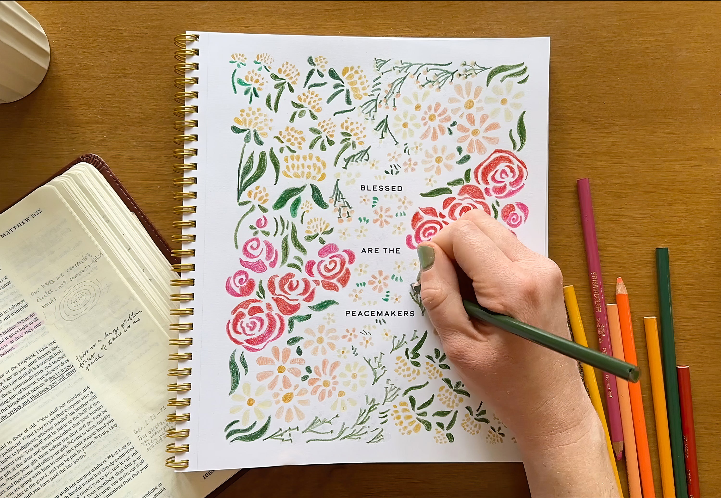 Person coloring a floral design in a spiral-bound notebook with colored pencils on a wooden table.