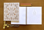 Open notebook with decorative cover on a wooden surface, accompanied by pencils and flowers.