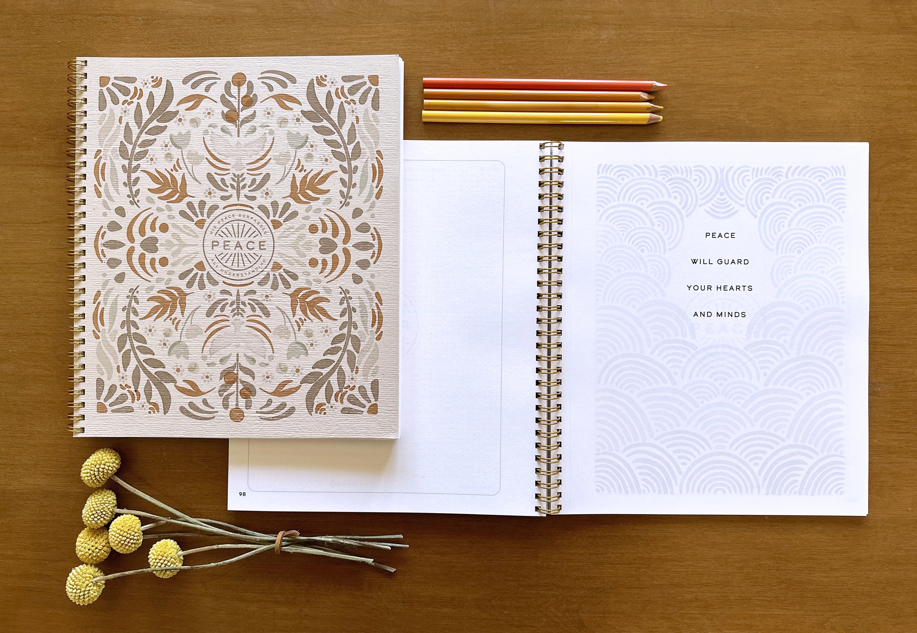 Open notebook with decorative cover on a wooden surface, accompanied by pencils and flowers.