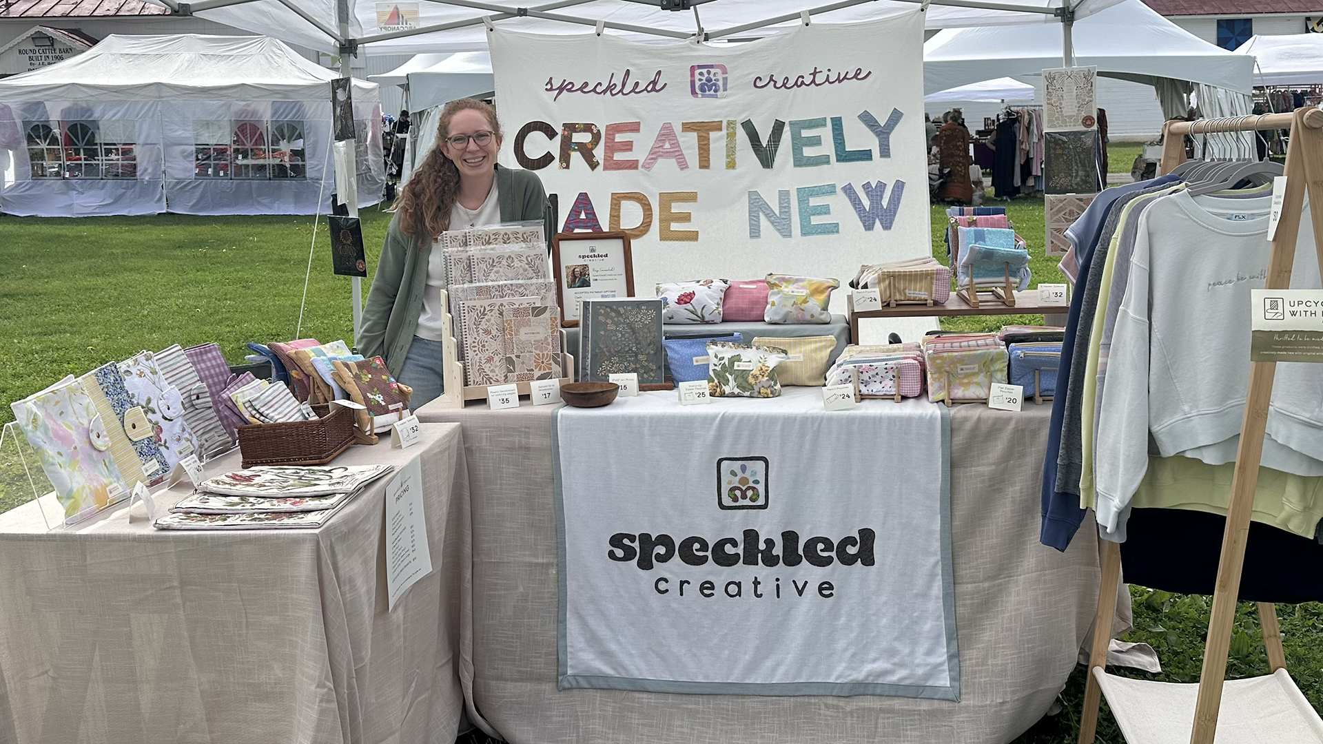 Woman standing at her market booth with handmade products and a banner that says Speckled Creative Creatively Made New