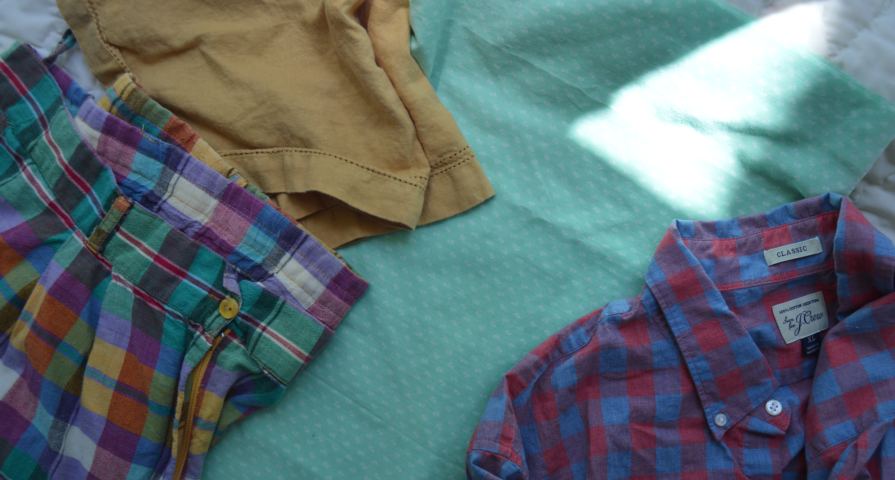 Three pieces of clothing including a plaid skirt, tan shorts, and a red and blue shirt on a green surface.
