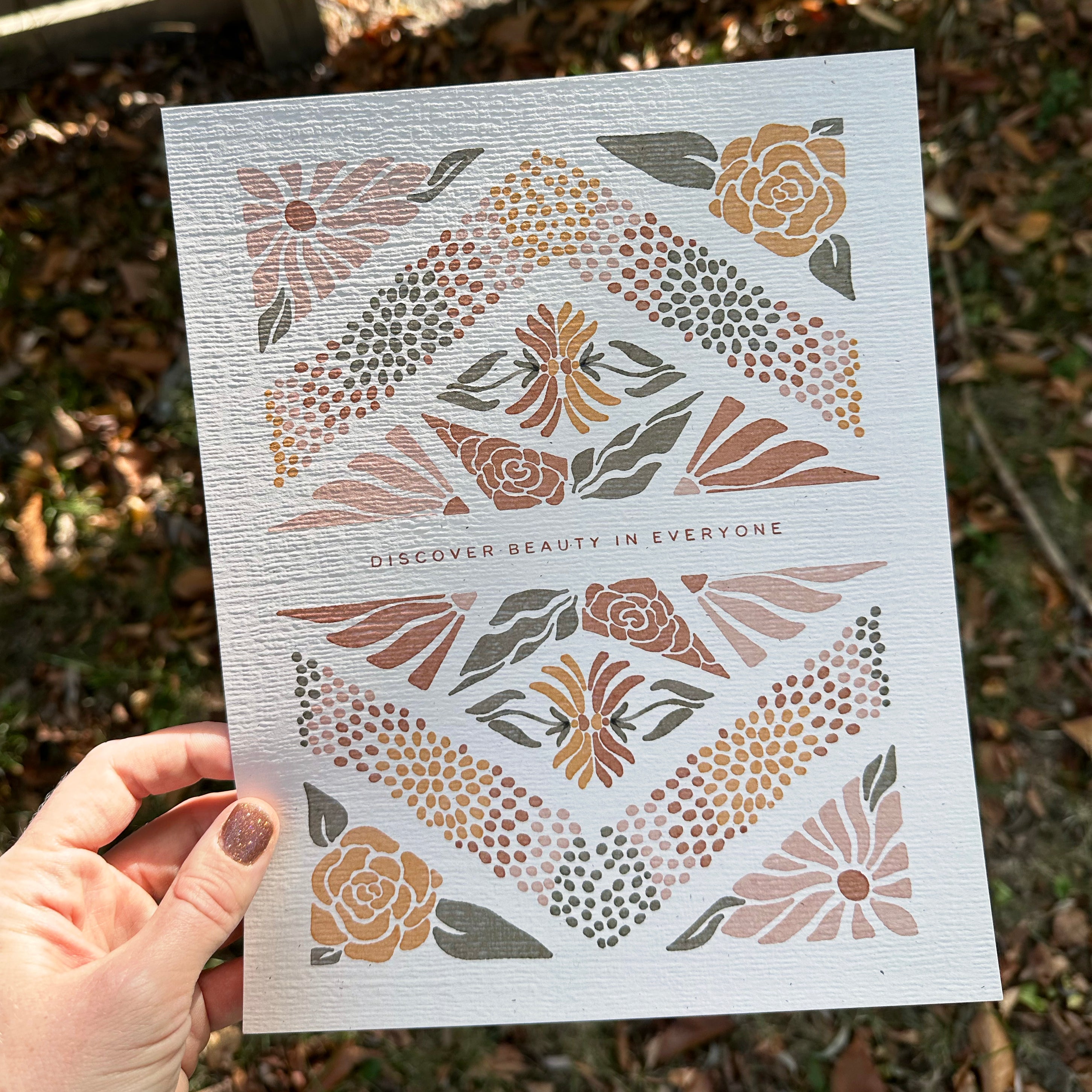 Hand holding art print with floral design and text that says "Discover the beauty in everyone" against a natural background.