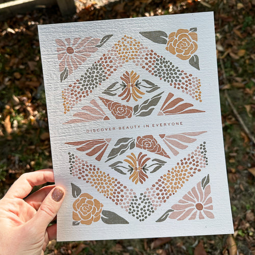 Hand holding art print with floral design and text that says "Discover the beauty in everyone" against a natural background.