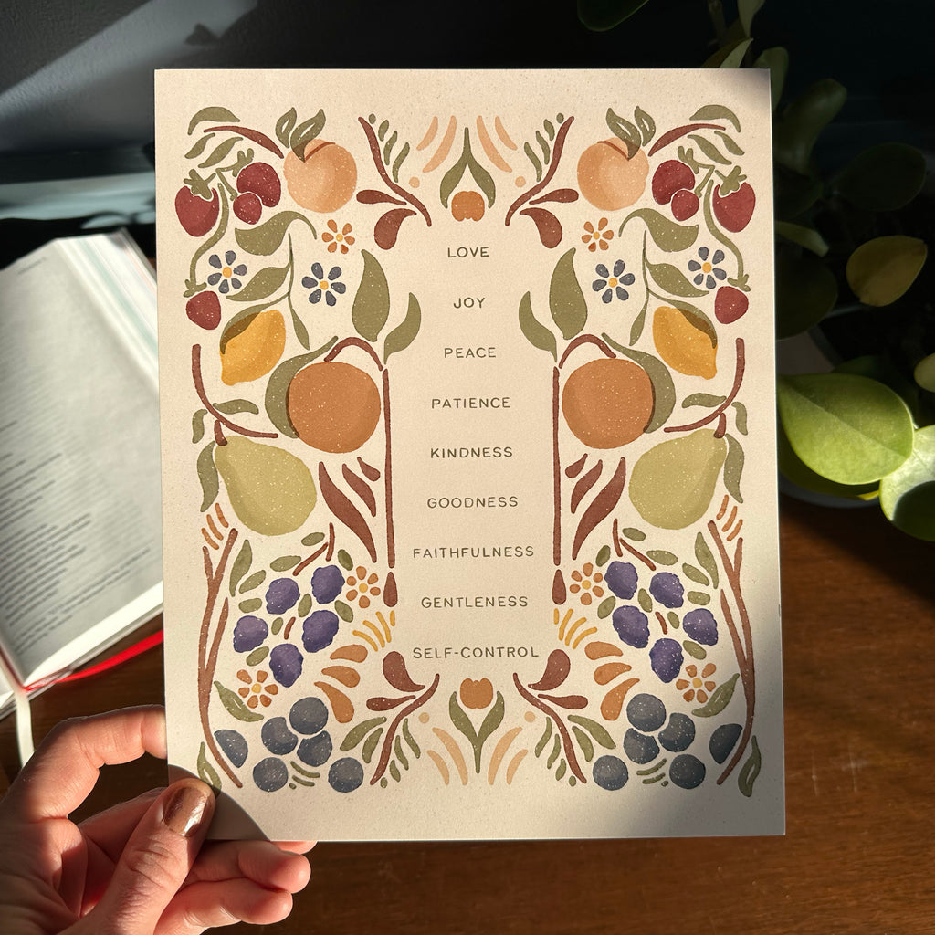 Hand holding art print with fruit illustrations that surround the words that describe the fruit of the Spirit