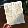 Hand holding art print with illustration of rocky terrain leading up to sheep and shepherd hooks surrounding the text "you will lie down and no one will make you afraid"