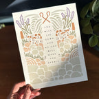 Hand holding art print with illustration of rocky terrain leading up to sheep and shepherd hooks surrounding the text "you will lie down and no one will make you afraid"