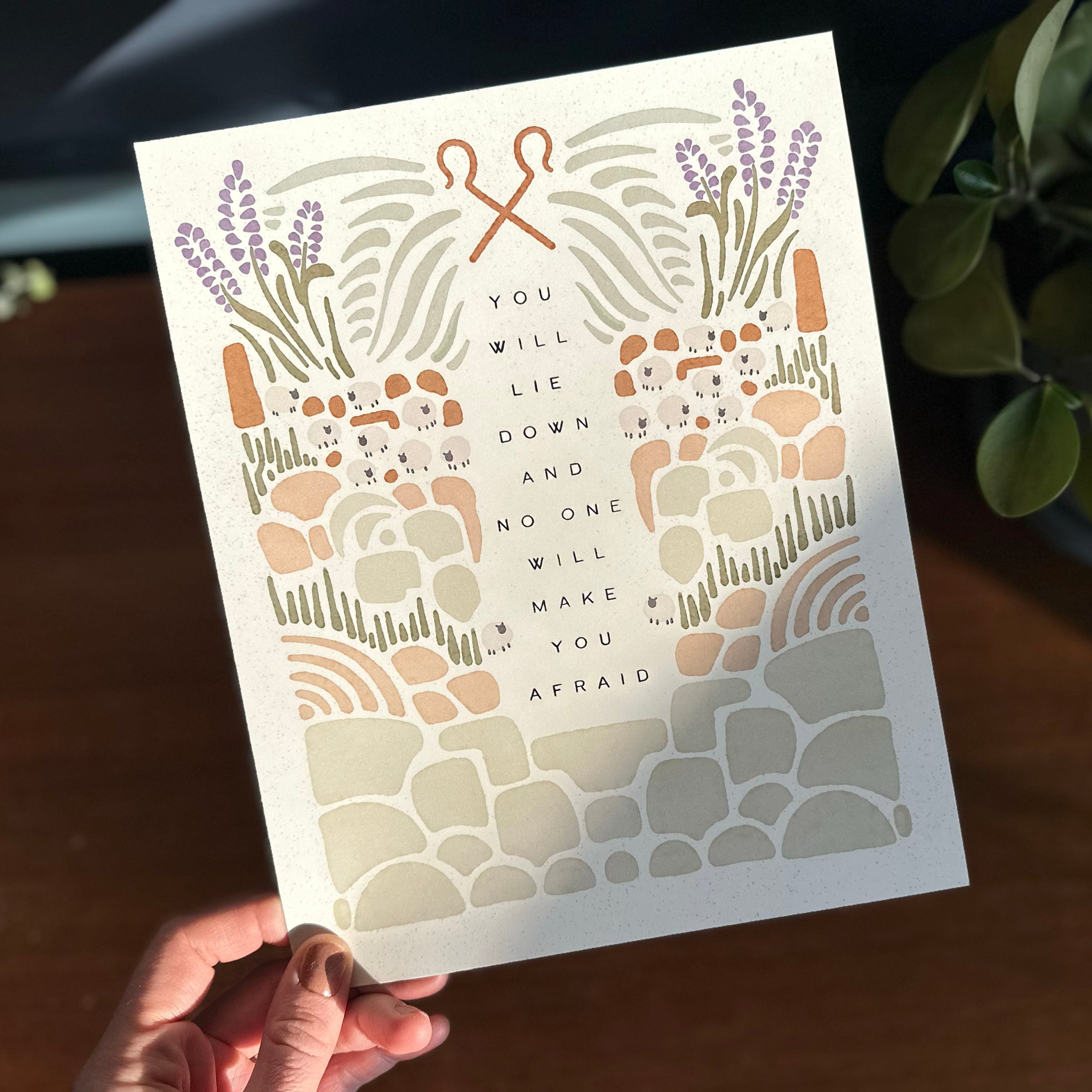 Hand holding art print with illustration of rocky terrain leading up to sheep and shepherd hooks surrounding the text "you will lie down and no one will make you afraid"