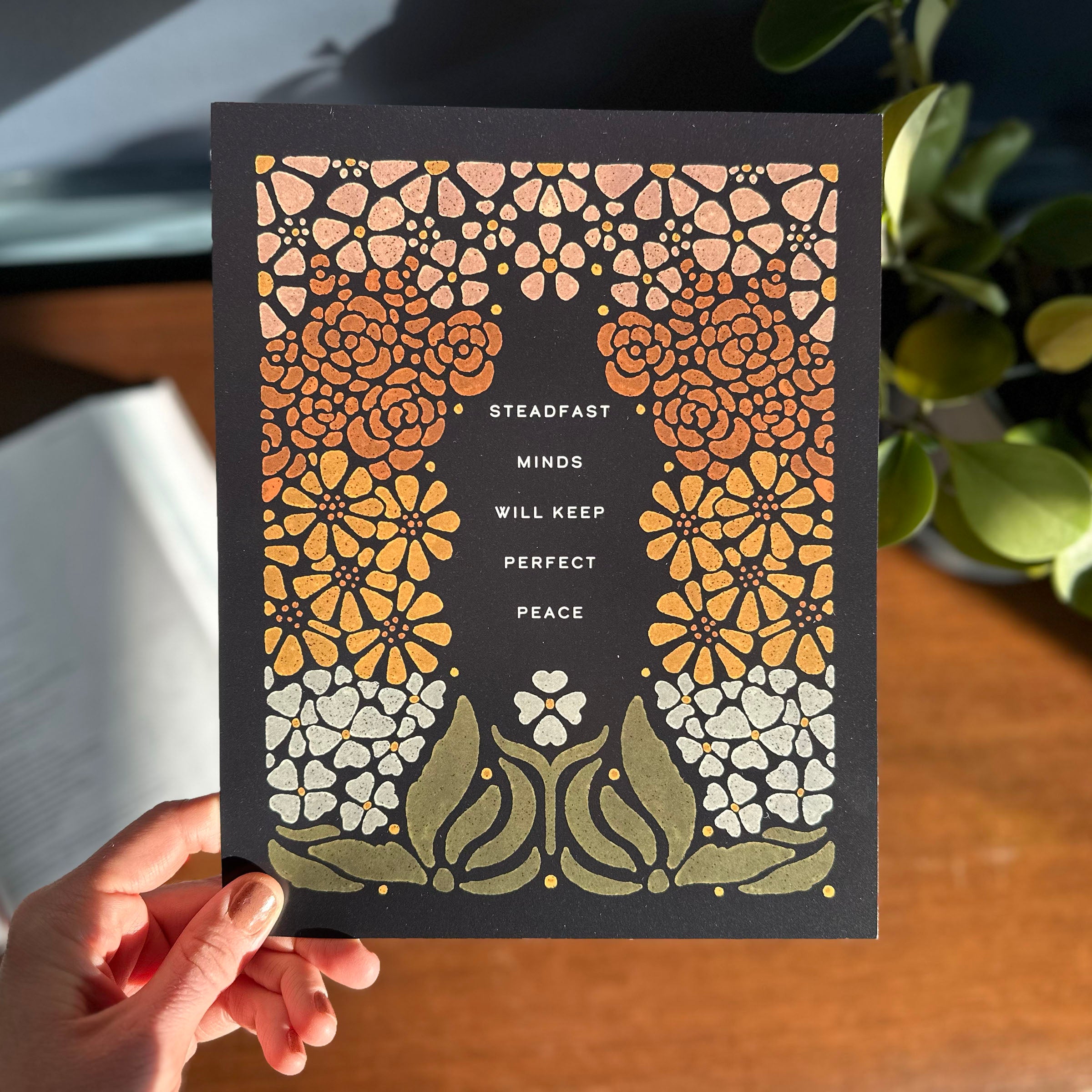 Hand holding art print with floral illustration surrounding words that say "steadfast minds will keep perfect peace"