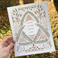 Hand holding an art print with decorative design and text that says "how good it is to dweel together in unity" against a natural background