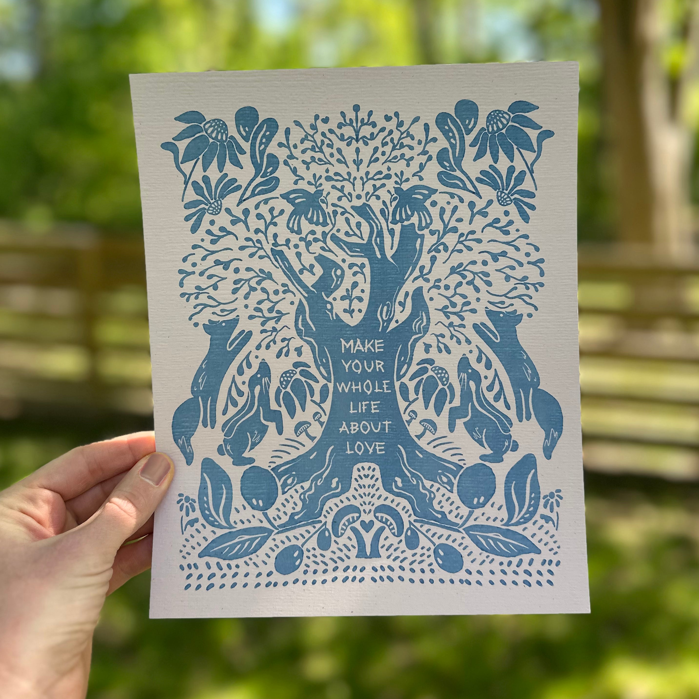 Art print with blue tree design and text that says "make your whole life about love" held by a hand outdoors