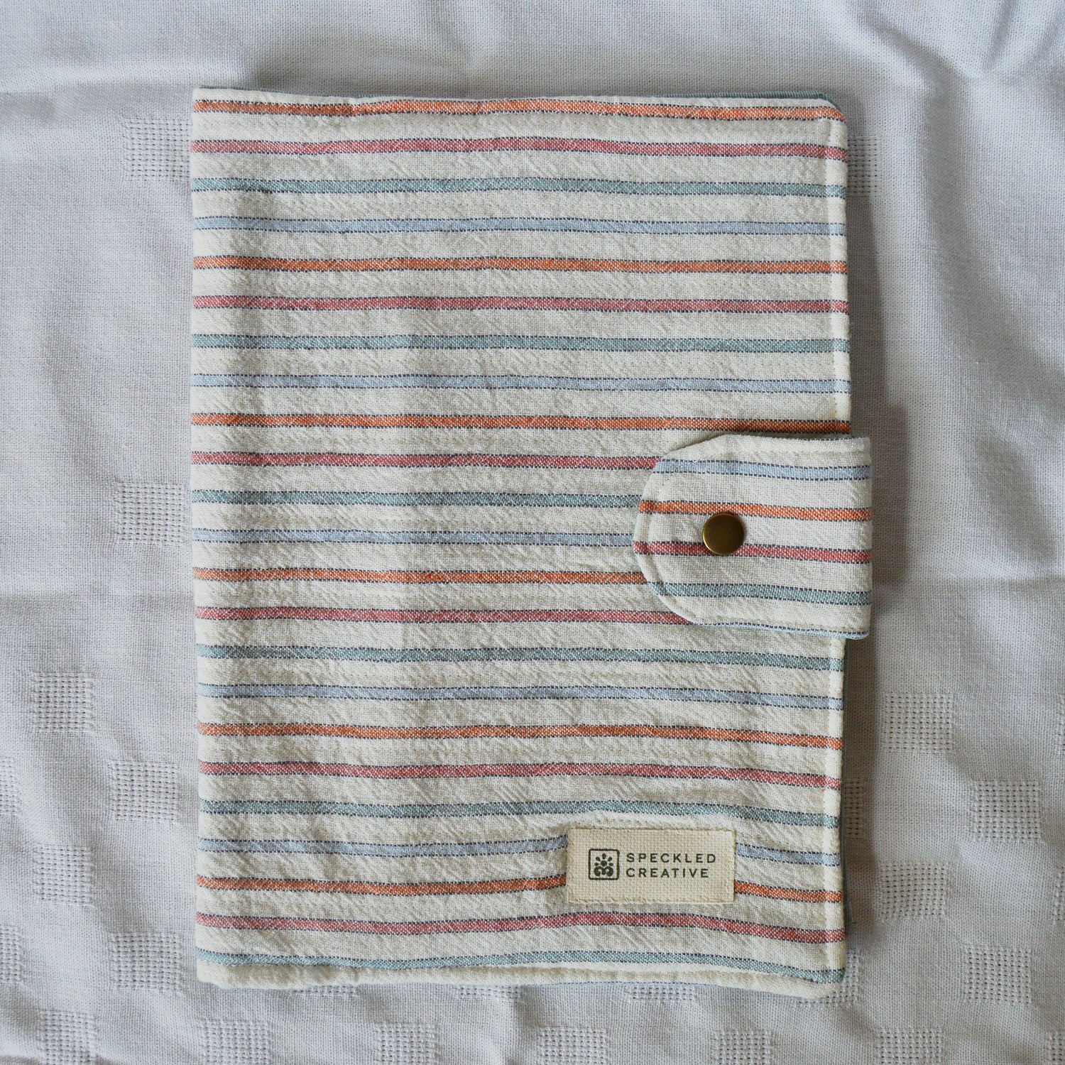 Striped fabric cover with a button closure on a textured surface, featuring the brand 'Speckled Creative'.