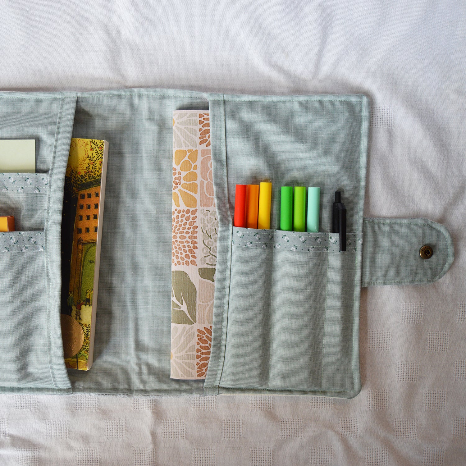 Teal fabric case with pockets containing books, a patterned book, and colorful markers on a white background.