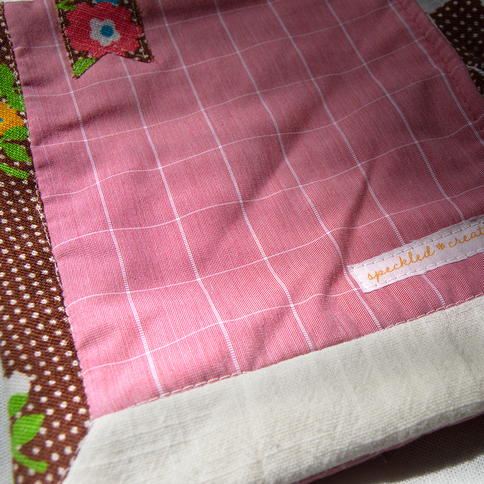 Close-up of a pink checkered fabric e-reader pouch  with a brand label on a textured surface.