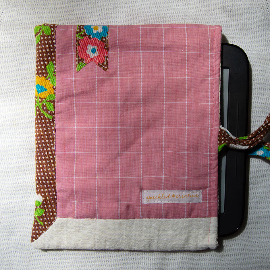 Fabric e-reader sleeve with pink grid pattern and floral accents covering a e-reader