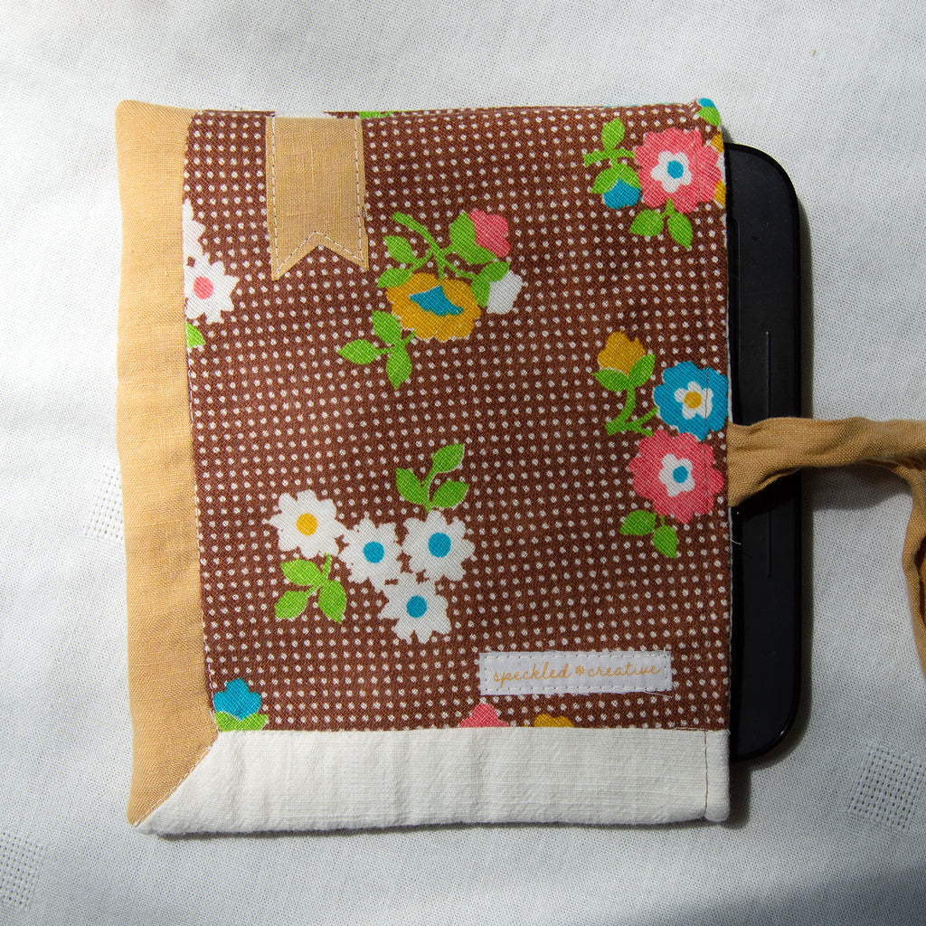 Floral-patterned fabric e-reader pouch with a visible brand label and a goldbookmark appliqué on a light gray background