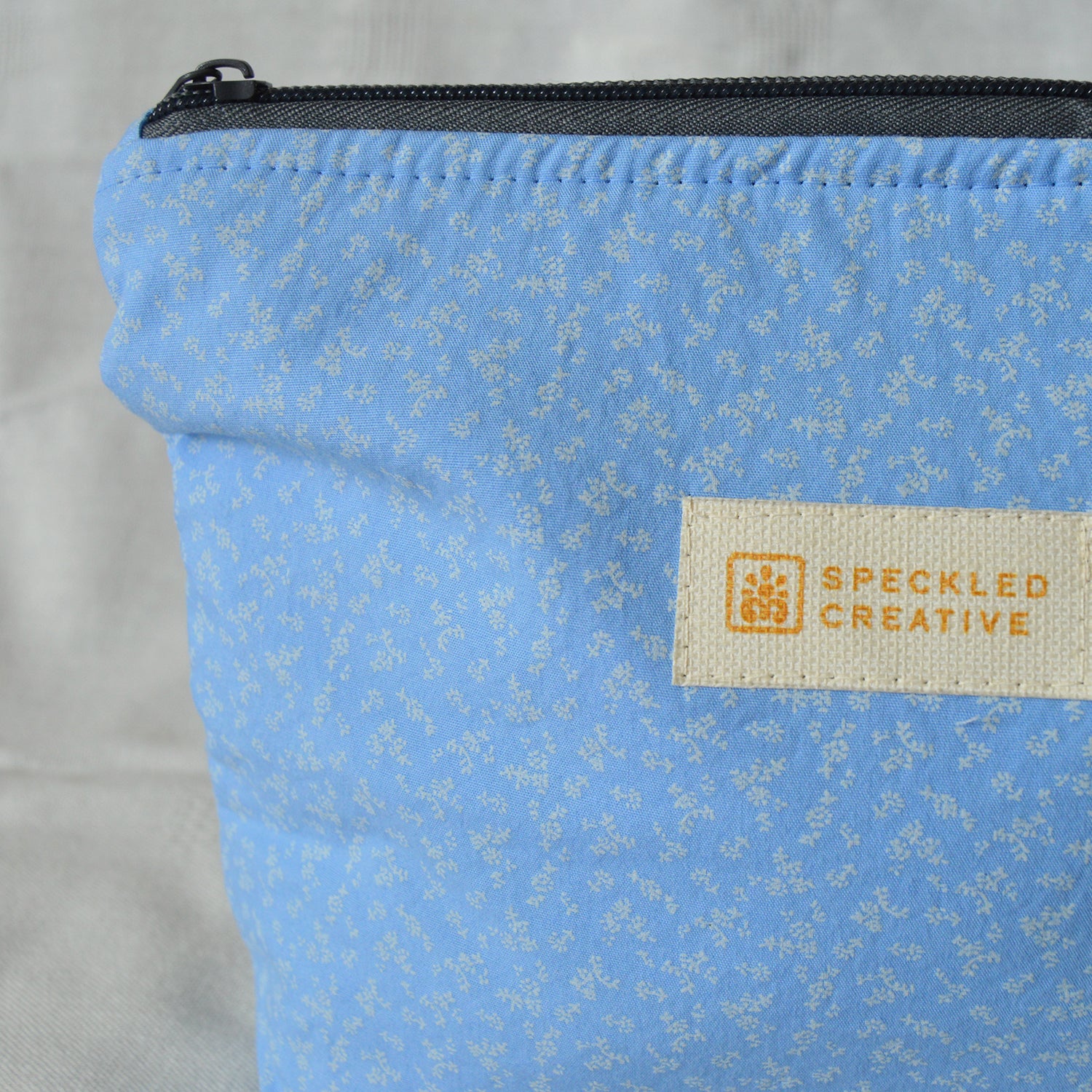 Close up of a blue pouch with a subtle fabric pattern and Speckled Creative label